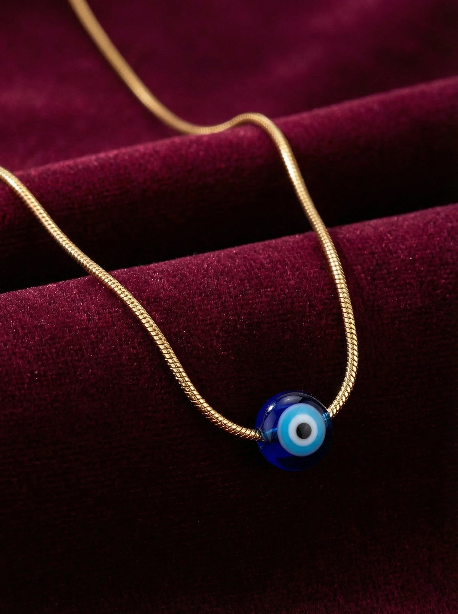 Gold necklace with a blue evil eye pendant on a dark red fabric background