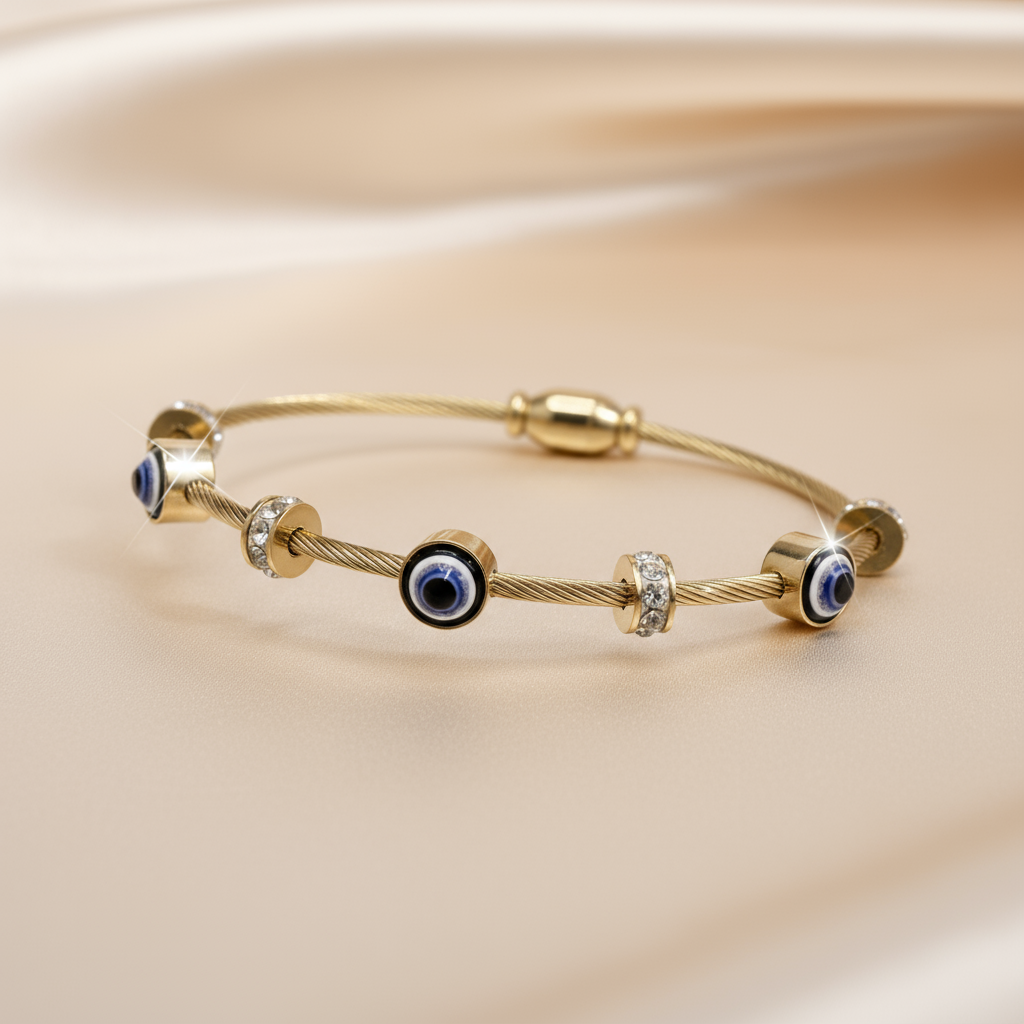 Studio product shot of the Gold-Tone Evil Eye Cable Wire Bangle, featuring crystal spacers, presented at a 45-degree angle on satin fabric.